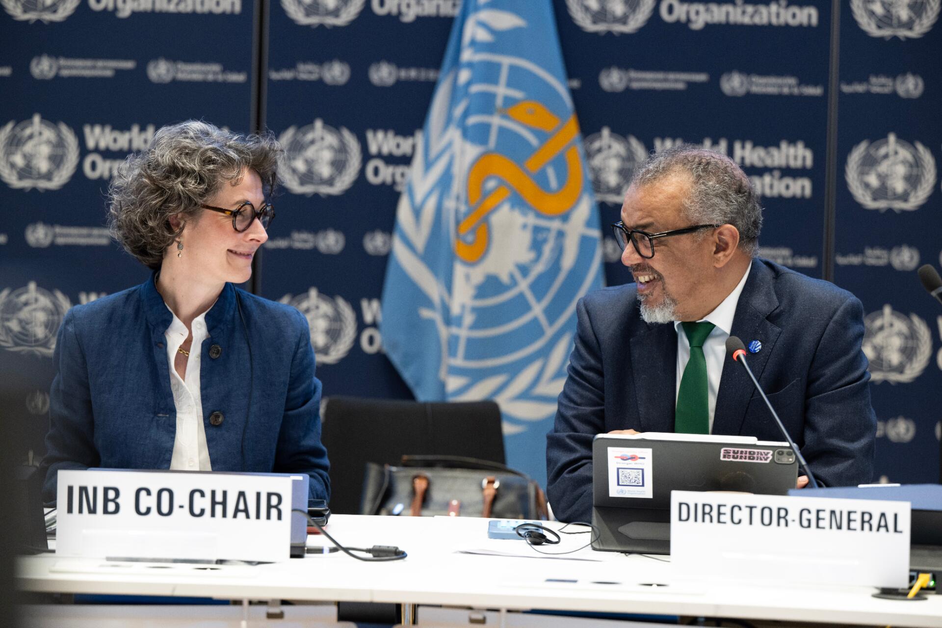 A woman and a man at a meeting podium, looking at each other smiling, with a WHO flag in the background.